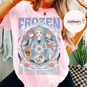 K&ouml;nnte beinhalten: Hellrosa T-Shirt mit einem farbenfrohen Grafikdesign aus dem Disney-Film Frozen. Das Design enth&auml;lt die W&ouml;rter "FROZEN" und "IN THIS TOGETHER" mit Bildern von Elsa, Anna, Kristoff, Olaf und Sven. Das T-Shirt hat einen Rundhalsausschnitt und kurze &Auml;rmel.