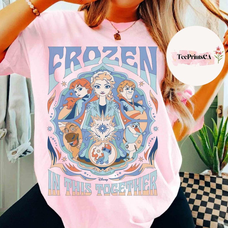 K&ouml;nnte beinhalten: Hellrosa T-Shirt mit einem farbenfrohen Grafikdesign aus dem Disney-Film Frozen. Das Design enth&auml;lt die W&ouml;rter "FROZEN" und "IN THIS TOGETHER" mit Bildern von Elsa, Anna, Kristoff, Olaf und Sven. Das T-Shirt hat einen Rundhalsausschnitt und kurze &Auml;rmel.