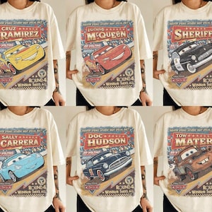 May include: A cream-colored t-shirt featuring a vintage-style graphic print of Disney Pixar Cars characters including Cruz Ramirez, Lightning McQueen, Sheriff, Sally Carrera, Doc Hudson, and Tow Mater with Radiator Springs race text.