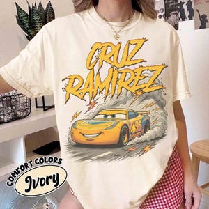 May include: Ivory-colored t-shirt featuring a cartoon image of a yellow race car with the text "CRUZ RAMIREZ" in yellow. The car has the number 95 on the side. The words "COMFORT COLORS" and "Ivory" are also printed on the shirt.