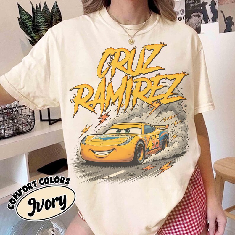 May include: Ivory-colored t-shirt featuring a cartoon image of a yellow race car with the text "CRUZ RAMIREZ" in yellow. The car has the number 95 on the side. The words "COMFORT COLORS" and "Ivory" are also printed on the shirt.
