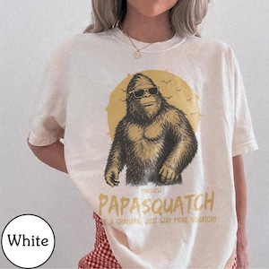May include: White t-shirt with a graphic of a Bigfoot wearing sunglasses in front of a yellow sun. The text reads "Papasquatch" and "Like a grandpa, just way more squatchy."