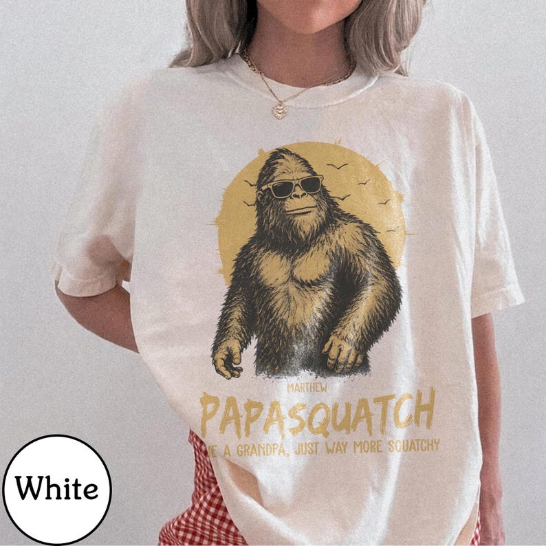 May include: White t-shirt with a graphic of a Bigfoot wearing sunglasses in front of a yellow sun. The text reads "Papasquatch" and "Like a grandpa, just way more squatchy."