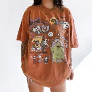 May include: An oversized burnt orange t-shirt featuring a graphic print of characters from The Nightmare Before Christmas, including Jack Skellington, Sally, Zero, and Oogie Boogie, along with various Halloween-themed icons and text elements.