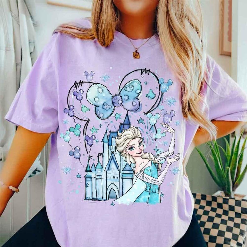 Pode incluir: Uma camiseta lil&aacute;s clara com uma estampa gr&aacute;fica de um castelo, uma princesa e um la&ccedil;o. O design incorpora elementos azuis, brancos e roxos. A camiseta tem mangas curtas e um corte folgado.