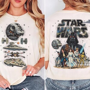 Könnte beinhalten: Cremefarbenes T-Shirt mit Star Wars-Grafiken. Die Vorderseite zeigt den Todesstern, Raumschiffe und den Millennium Falken. Die Rückseite zeigt Darth Vader, Charaktere und das Star Wars-Logo.