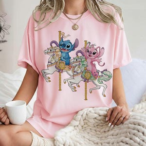 May include: Pink t-shirt featuring a carousel scene with blue and pink cartoon characters riding white horses. The carousel has gold poles and decorative elements. The shirt is a solid pink color.