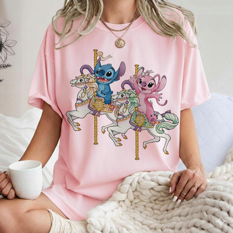 May include: Pink t-shirt featuring a carousel scene with blue and pink cartoon characters riding white horses. The carousel has gold poles and decorative elements. The shirt is a solid pink color.