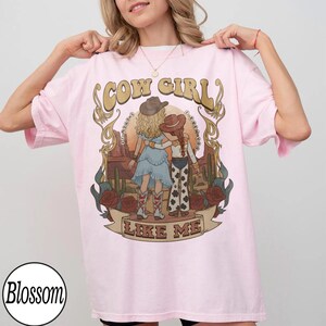 Może przedstawiać: Jasnor&oacute;żowy t-shirt z grafiką przedstawiającą dwie postacie w kowbojskich kapeluszach na tle pustynnego krajobrazu z kaktusami i r&oacute;żami. Napis "COW GIRL" znajduje się nad postaciami, a "LIKE ME" jest wydrukowany na wstędze poniżej.