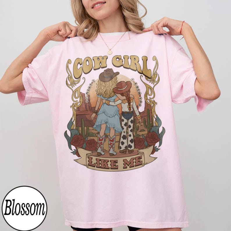 Może przedstawiać: Jasnor&oacute;żowy t-shirt z grafiką przedstawiającą dwie postacie w kowbojskich kapeluszach na tle pustynnego krajobrazu z kaktusami i r&oacute;żami. Napis "COW GIRL" znajduje się nad postaciami, a "LIKE ME" jest wydrukowany na wstędze poniżej.