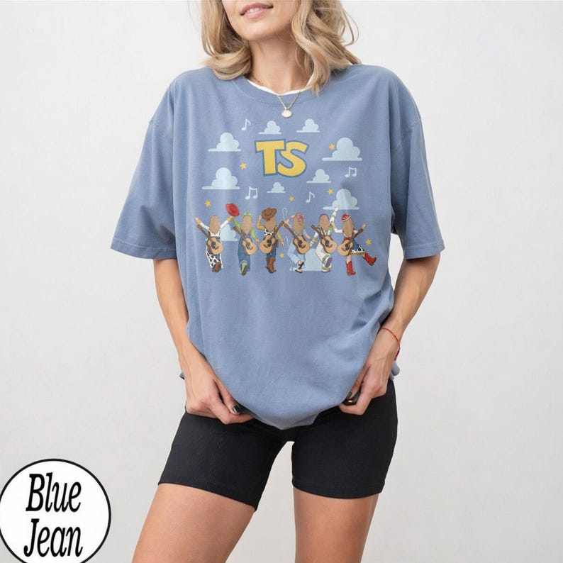 May include: A blue denim-colored t-shirt featuring a graphic print of six stylized figures playing acoustic guitars. Above the figures, the letters TS are printed in yellow, surrounded by white cloud shapes, musical notes, and small yellow stars.