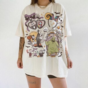 May include: An off-white short-sleeve t-shirt featuring a colorful graphic print of characters and icons from The Nightmare Before Christmas, including Jack Skellington, Sally, Oogie Boogie, and Zero, with the text Nightmare arranged in a circle.