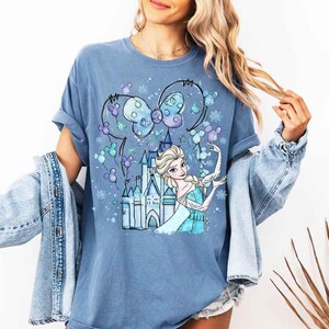 Pode incluir: Camiseta azul com uma ilustra&ccedil;&atilde;o de desenho animado de uma princesa em um vestido azul, um castelo e um la&ccedil;o com orelhas de rato. O design inclui flocos de neve e outros elementos decorativos. A camiseta &eacute; azul lisa.