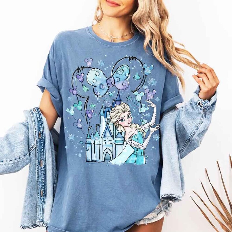 Pode incluir: Camiseta azul com uma ilustra&ccedil;&atilde;o de desenho animado de uma princesa em um vestido azul, um castelo e um la&ccedil;o com orelhas de rato. O design inclui flocos de neve e outros elementos decorativos. A camiseta &eacute; azul lisa.