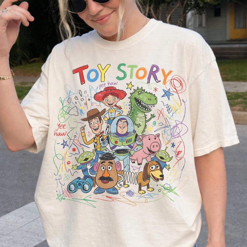May include: Cream-colored t-shirt featuring a colorful graphic of Toy Story characters, including Woody, Buzz Lightyear, and Rex. The words "Toy Story" are written in a playful, multi-colored font above the characters. The shirt has a crew neck and short sleeves.