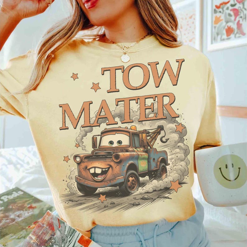 May include: A light yellow t-shirt featuring the text "TOW MATER" in orange letters with a cartoon image of Mater the tow truck. The design includes stars and a cloud of smoke, creating a vintage, animated style.