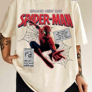 Op de afbeelding: Ivoor T-shirt met een Spider-Man afbeelding. Het ontwerp bevat de tekst "BRAND NEW DAY SPIDER-MAN" in rood en blauw, stripstijlelementen en de tekst "ANOTHER SPECTACULAR SMASH-HIT FROM THE HOUSE OF IDEAS!"