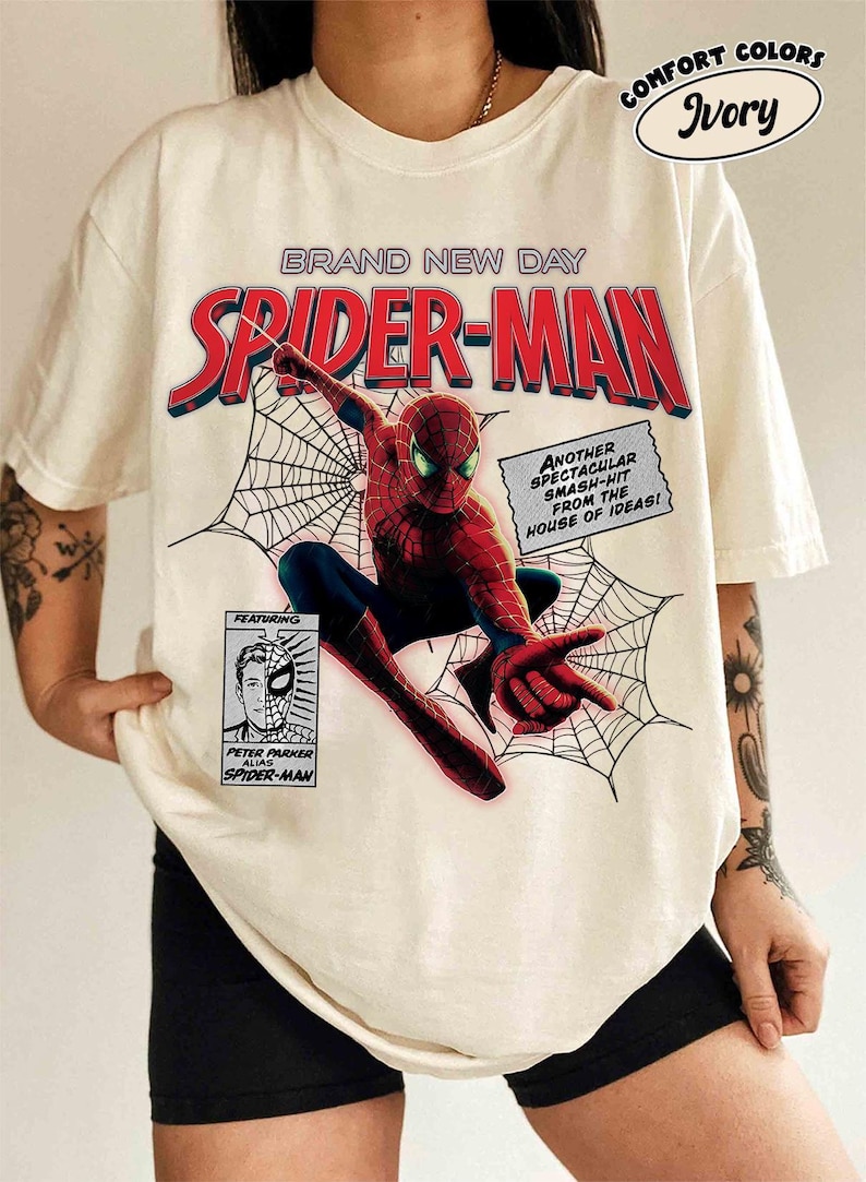 Op de afbeelding: Ivoor T-shirt met een Spider-Man afbeelding. Het ontwerp bevat de tekst "BRAND NEW DAY SPIDER-MAN" in rood en blauw, stripstijlelementen en de tekst "ANOTHER SPECTACULAR SMASH-HIT FROM THE HOUSE OF IDEAS!"