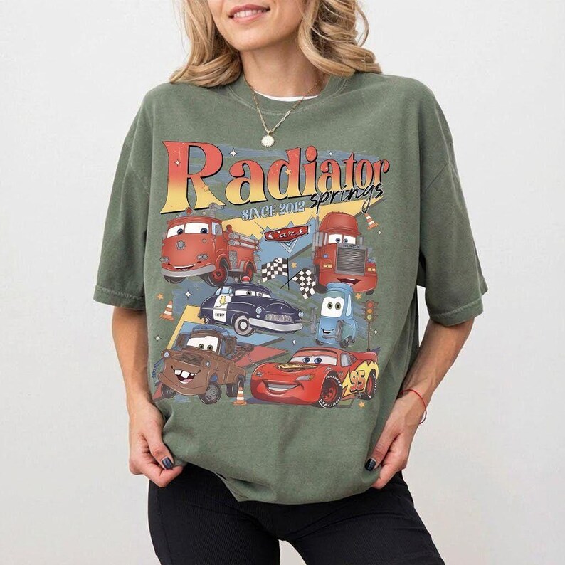 May include: Green t-shirt featuring a graphic of characters from the movie Cars, including Lightning McQueen and Mater. The design includes the text "Radiator Springs Since 2012" in a retro style. The shirt has short sleeves and a relaxed fit.