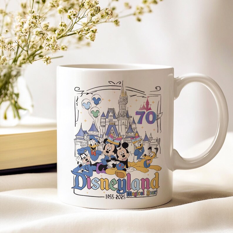 May include: White ceramic mug featuring a Disneyland design. The mug showcases Mickey Mouse, Minnie Mouse, Donald Duck, and Pluto in front of a castle. The mug has the text "Disneyland" and "1955-2025 Magical Year" with the number 70.