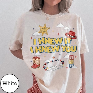 May include: A white t-shirt featuring a graphic print with the text I KNEW IT I KNEW YOU. The design includes a yellow sheriff star badge, a red cowboy hat, a pair of red cowboy boots, a beaded bracelet with the text YOU'VE GOT A FRIEND IN ME, and stacked alphabet blocks labeled T, S, and S, surrounded by small clouds and stars.