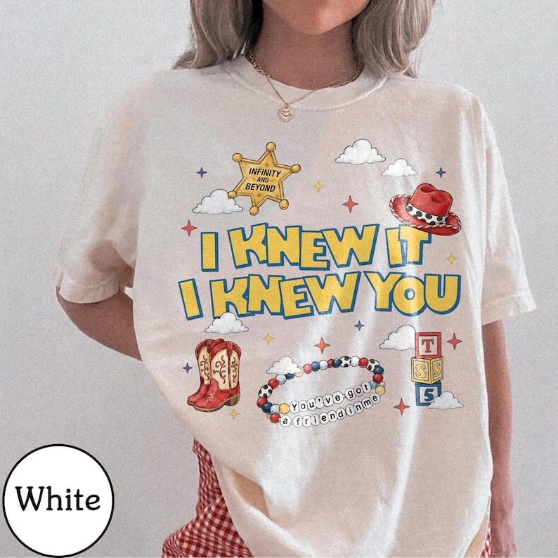 May include: A white t-shirt featuring a graphic print with the text I KNEW IT I KNEW YOU. The design includes a yellow sheriff star badge, a red cowboy hat, a pair of red cowboy boots, a beaded bracelet with the text YOU'VE GOT A FRIEND IN ME, and stacked alphabet blocks labeled T, S, and S, surrounded by small clouds and stars.