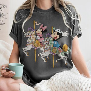 May include: Dark gray t-shirt featuring a carousel scene with Mickey Mouse, Minnie Mouse, and a carousel horse design. The carousel horses are white with colorful accents. Mickey Mouse is wearing a blue outfit and Minnie Mouse is wearing a pink dress.