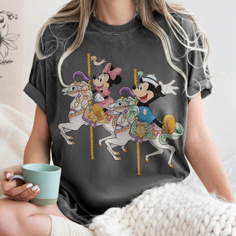 May include: Dark gray t-shirt featuring a carousel scene with Mickey Mouse, Minnie Mouse, and a carousel horse design. The carousel horses are white with colorful accents. Mickey Mouse is wearing a blue outfit and Minnie Mouse is wearing a pink dress.