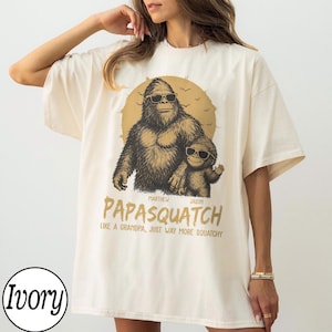 May include: Ivory-colored t-shirt featuring a graphic of a Bigfoot-like creature and a smaller version, both wearing sunglasses. The text "PAPASQUATCH" is below the image, with the tagline "LIKE A GRANDPA, JUST WAY MORE SQUATCHY."