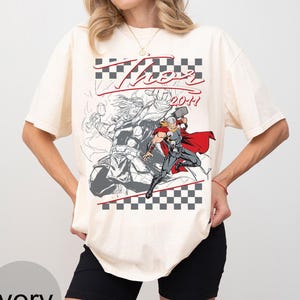 May include: Ivory-colored t-shirt featuring a graphic of Thor, with the word "Thor" in red script and the year "2011". The design includes a checkered pattern and a comic-style illustration of the superhero.