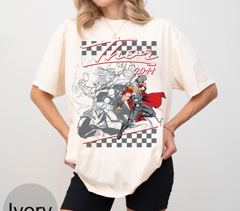 May include: Ivory-colored t-shirt featuring a graphic of Thor, with the word "Thor" in red script and the year "2011". The design includes a checkered pattern and a comic-style illustration of the superhero.
