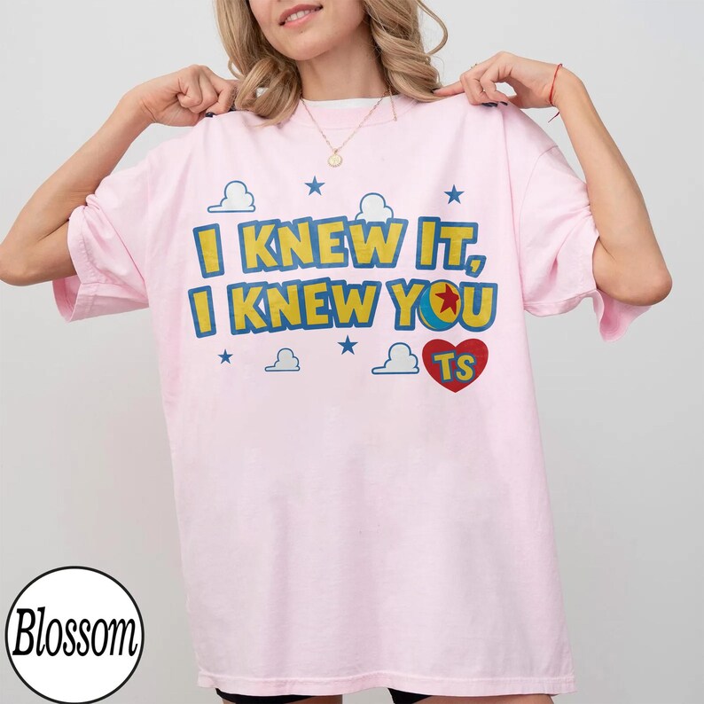 Pu&ograve; includere: Una t-shirt a maniche corte rosa chiaro con il testo giallo I KNEW IT, I KNEW YOU e un cuore rosso contenente le lettere TS. Il design include stelle blu e grafiche di nuvole bianche stampate sulla parte anteriore del capo.