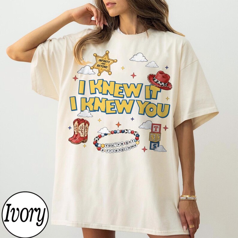 May include: An ivory t-shirt featuring a graphic print with the text I KNEW IT I KNEW YOU in yellow and blue. The design includes a sheriff badge, a red cowboy hat, red cowboy boots, a beaded bracelet with the text YOU'VE GOT A FRIEND IN ME, and blocks.