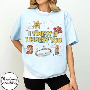 May include: A light blue t-shirt featuring a graphic print with the text I KNEW IT I KNEW YOU, a gold sheriff badge with INFINITY AND BEYOND, a red cowboy hat, cowboy boots, a beaded bracelet with YOU'VE GOT A FRIEND IN ME, and toy blocks.
