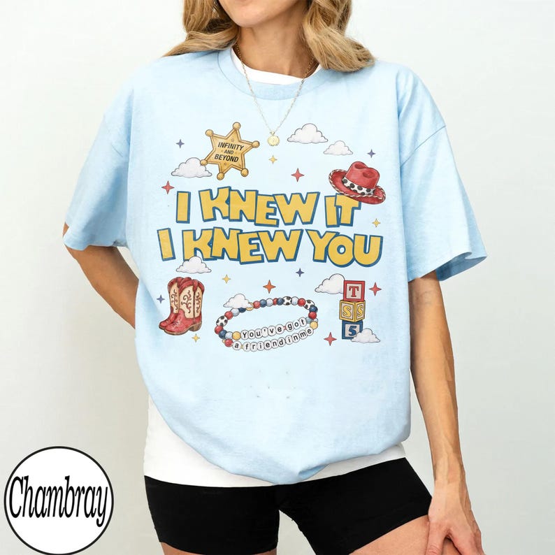 May include: A light blue t-shirt featuring a graphic print with the text I KNEW IT I KNEW YOU, a gold sheriff badge with INFINITY AND BEYOND, a red cowboy hat, cowboy boots, a beaded bracelet with YOU'VE GOT A FRIEND IN ME, and toy blocks.