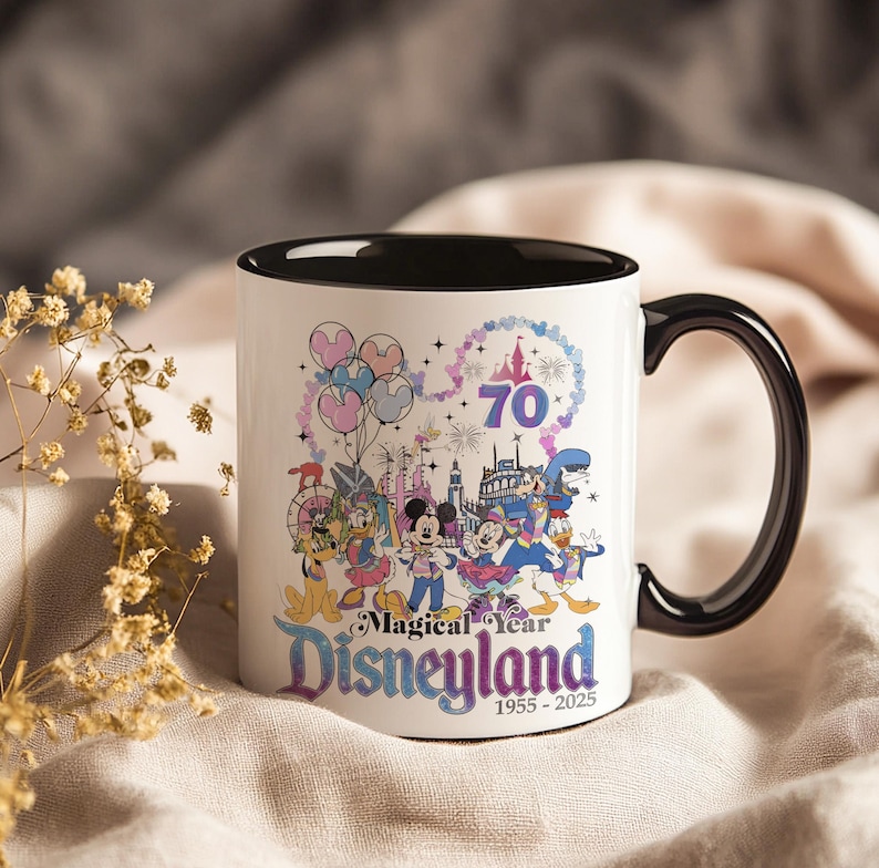 Puede incluir: Taza de cer&aacute;mica blanca con interior y asa negros. La taza presenta un gr&aacute;fico colorido de personajes de Disney, fuegos artificiales y el texto "Magical Year Disneyland 1955-2025" celebrando su 70 aniversario.