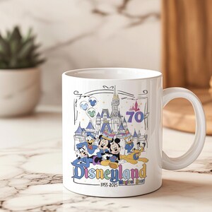 May include: White ceramic mug featuring a Disneyland design. The design includes Mickey Mouse, Donald Duck, Goofy, and a castle, with the text "Disneyland" and "1955-2025 Magical Year". The mug has a curved handle and is suitable for hot or cold beverages.