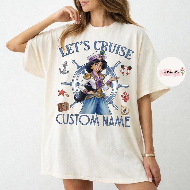 Pu&ograve; includere: T-shirt color crema con grafica di una donna in uniforme da capitano, circondata da elementi nautici. Il testo "LET'S CRUISE" e "CUSTOM NAME" sono stampati in blu. Include un'ancora, una stella marina e una bussola.