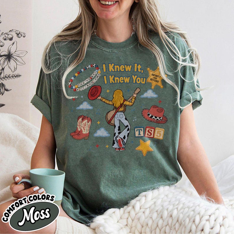 May include: A moss green short-sleeve t-shirt featuring a graphic print with the text I Knew It, I Knew You, Infinity and Beyond, and TS5. The design includes a person with blonde hair, a guitar, cowboy boots, hats, and a beaded bracelet.