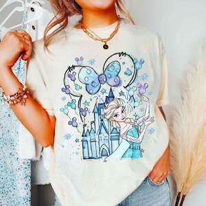 Pode incluir: Camiseta creme com uma ilustra&ccedil;&atilde;o de desenho animado de uma princesa em um vestido azul, um castelo e um la&ccedil;o com orelhas de rato. O design incorpora detalhes em azul, roxo e branco. A camiseta &eacute; uma pe&ccedil;a de roupa casual.