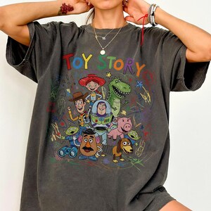 May include: Dark gray t-shirt featuring a colorful graphic of characters from the animated film "Toy Story." The design includes Woody, Buzz Lightyear, Rex, and other characters, with the words "Toy Story" in a playful font.