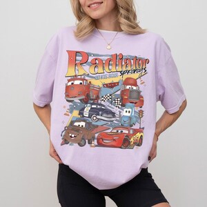May include: A light purple t-shirt featuring a graphic of various cartoon cars with the text "Radiator Springs Since 2012". The design includes a fire truck, police car, and race cars.