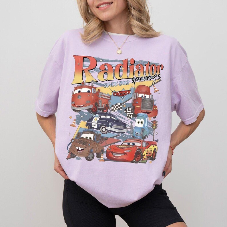 May include: A light purple t-shirt featuring a graphic of various cartoon cars with the text "Radiator Springs Since 2012". The design includes a fire truck, police car, and race cars.