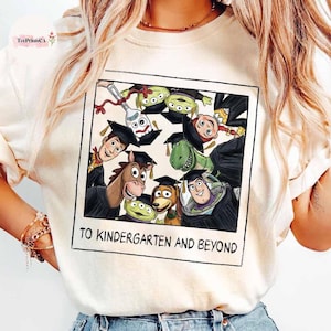 Puede incluir: Sudadera color crema con un gráfico enmarcado de personajes de Toy Story con gorros de graduación. La imagen incluye a Woody, Buzz Lightyear y otros. El texto "TO KINDERGARTEN AND BEYOND" está impreso debajo.