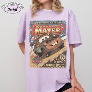 May include: A light purple t-shirt featuring a graphic print of the character Tow Mater from the movie Cars. The design includes the text Radiator Springs Saturday Night Special Race, Tow Mater, and Round 4 Winner Takes All, with a checkered pattern.