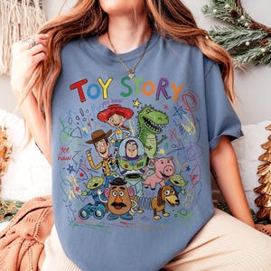 May include: A light blue t-shirt featuring the words "TOY STORY" in colorful letters. The shirt showcases a vibrant graphic of characters from the animated film, including Woody, Buzz Lightyear, and Rex, with scribbled accents.