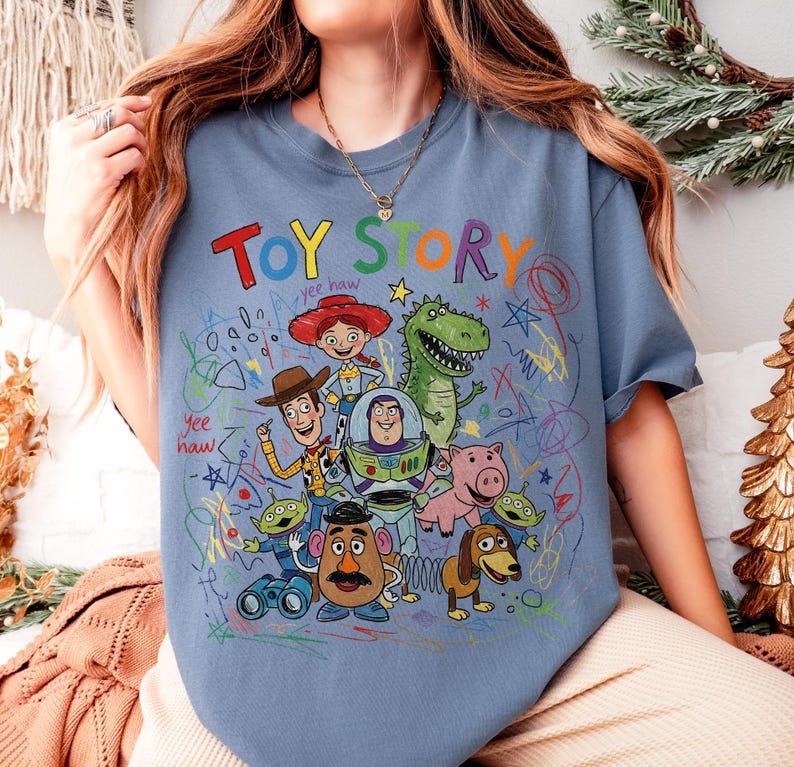 May include: A light blue t-shirt featuring the words "TOY STORY" in colorful letters. The shirt showcases a vibrant graphic of characters from the animated film, including Woody, Buzz Lightyear, and Rex, with scribbled accents.