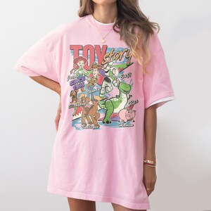 Puede incluir: Camiseta rosa claro con un dise&ntilde;o de personajes de Toy Story, incluyendo a Woody, Buzz Lightyear y Jessie. El dise&ntilde;o incluye el texto "Toy Story" y "Since 1995". La camiseta tiene ribetes blancos en las mangas.