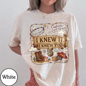 May include: A white t-shirt featuring a graphic print with a brown scroll banner that reads I KNEW IT, I KNEW YOU. The design includes a large number 5, brown cowboy boots, a red cowboy hat, alphabet blocks, a beaded necklace, and the text Cinfinity and beyond.