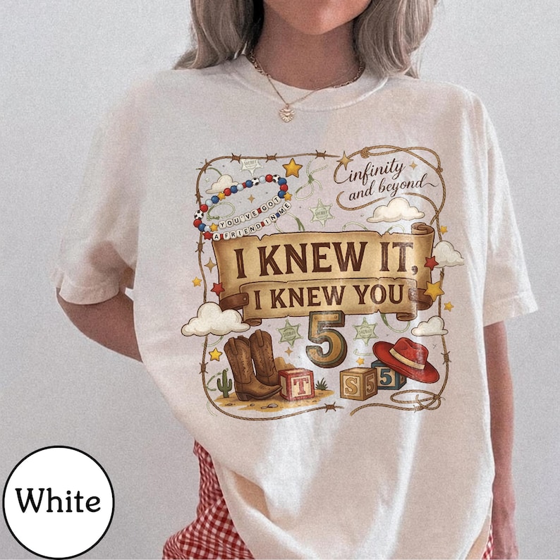 May include: A white t-shirt featuring a graphic print with a brown scroll banner that reads I KNEW IT, I KNEW YOU. The design includes a large number 5, brown cowboy boots, a red cowboy hat, alphabet blocks, a beaded necklace, and the text Cinfinity and beyond.